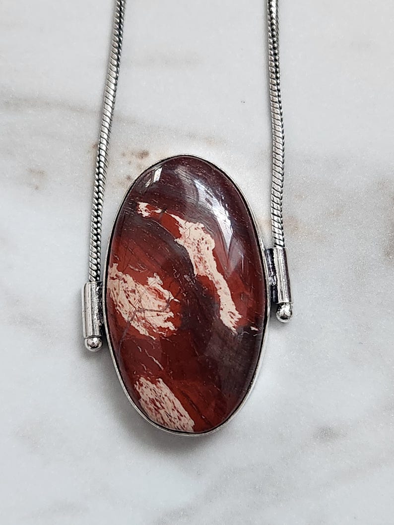 Peut inclure: Un collier argent&eacute; avec un pendentif ovale. Le pendentif pr&eacute;sente une pierre polie brun-rouge&acirc;tre avec des veines blanches et cr&egrave;me. La pierre est sertie dans une lunette argent&eacute;e et suspendue &agrave; une cha&icirc;ne argent&eacute;e.