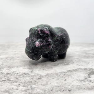 May include: A small, hand-carved figurine of a hippopotamus crafted from a dark, mottled stone. The mineral features a deep black base color with prominent, irregular patches of vibrant magenta and dark green throughout its polished surface.