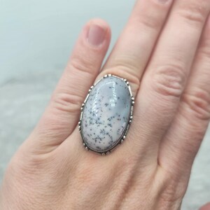 May include: Oval-shaped ring featuring a large, light gray stone with black speckles, set in a silver-toned metal band. The ring has a decorative, beaded edge. The ring is worn on a hand.