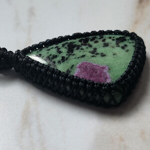 Peut inclure: Un pendentif en forme de goutte compos&eacute; d'une pierre verte polie avec des taches noires et une inclusion magenta pro&eacute;minente. La pierre est ench&acirc;ss&eacute;e dans un sertissage en cordon macram&eacute; noir &agrave; la texture tress&eacute;e, fix&eacute; &agrave; un cordon noir.