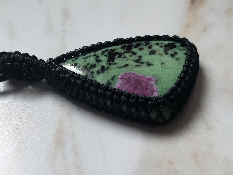Peut inclure: Un pendentif en forme de goutte compos&eacute; d'une pierre verte polie avec des taches noires et une inclusion magenta pro&eacute;minente. La pierre est ench&acirc;ss&eacute;e dans un sertissage en cordon macram&eacute; noir &agrave; la texture tress&eacute;e, fix&eacute; &agrave; un cordon noir.