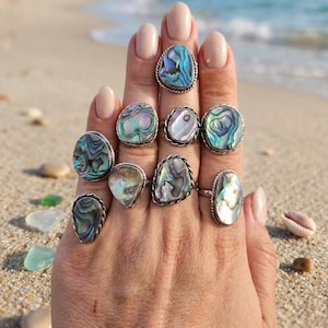 May include: A hand displays ten silver rings with iridescent abalone shells. The rings have various shapes, including oval, round, and teardrop. The abalone shells display shades of blue, green, and purple. The background is a sandy beach with the ocean in the distance.