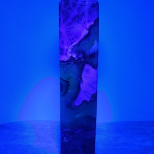 May include: A tall, rectangular crystal obelisk with a pointed top. The stone features a mix of dark blue, green, and purple hues, with a marbled pattern. The obelisk is set against a solid blue background.