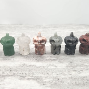 May include: Eight small carved pumpkin figurines made of various types of stone. The pumpkins are all in a row and are facing forward. The pumpkins are in the following colors: black, green, white, brown, gray, black, red, and black.