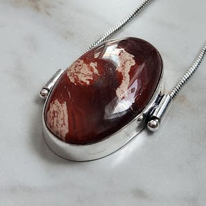 Peut inclure: Un collier en argent avec un pendentif ovale. Le pendentif pr&eacute;sente une pierre brun-rouge polie avec des marbrures cr&egrave;me, sertie dans une lunette en argent. La cha&icirc;ne est &eacute;galement en argent.
