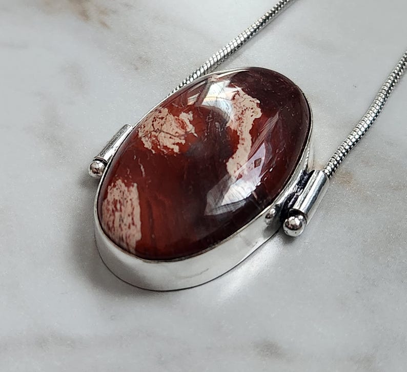 Peut inclure: Un collier en argent avec un pendentif ovale. Le pendentif pr&eacute;sente une pierre brun-rouge polie avec des marbrures cr&egrave;me, sertie dans une lunette en argent. La cha&icirc;ne est &eacute;galement en argent.