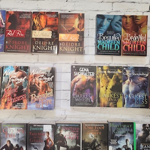 May include: A collection of paperback romance novels with various titles and authors. The books are arranged in rows on a white brick wall background.