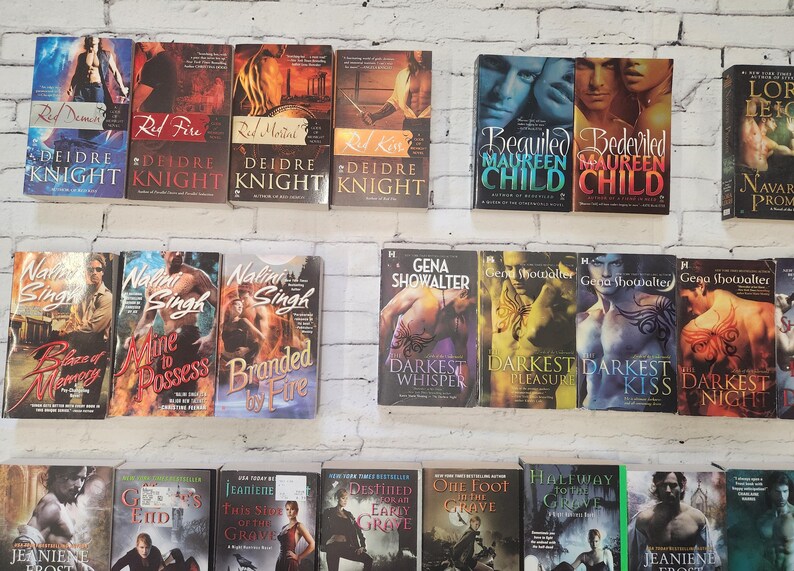 May include: A collection of paperback romance novels with various titles and authors. The books are arranged in rows on a white brick wall background.
