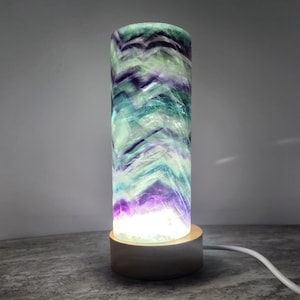 May include: A cylindrical lamp made of natural fluorite stone featuring bands of purple, teal, and white in a chevron pattern. The stone is illuminated from within by a light source and is mounted on a circular, light-colored wooden base.