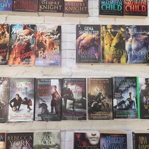 May include: A shelf of paperback books with various titles and authors. The books are arranged in rows and are mostly romance novels. Some of the titles include "Blazed" by Nalini Singh, "Mine to Possess" by Nalini Singh, "Branded by Fire" by Nalini Singh, "Darkest Whisper" by Gena Showalter, "Darkest Pleasure" by Gena Showalter, "Darkest Kiss" by Gena Showalter, "Darkest Night" by Gena Showalter, "Wicked Night" by Jeaniene Frost, "Ghost's End" by Jeaniene Frost, "This Side of the Grave" by Jeaniene Frost, "Destined for an Early Grave" by Jeaniene Frost, "One Foot in the Grave" by Jeaniene Frost, "Halfway to the Grave" by Jeaniene Frost, "Wicked Bite" by Jeaniene Frost, "Frost Bound" by Jeaniene Frost.