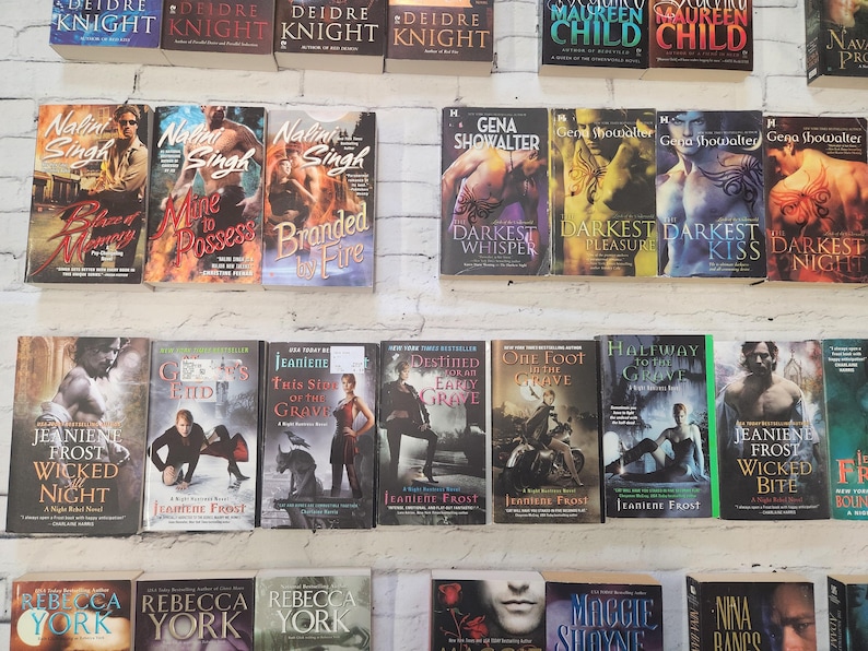 May include: A shelf of paperback books with various titles and authors. The books are arranged in rows and are mostly romance novels. Some of the titles include "Blazed" by Nalini Singh, "Mine to Possess" by Nalini Singh, "Branded by Fire" by Nalini Singh, "Darkest Whisper" by Gena Showalter, "Darkest Pleasure" by Gena Showalter, "Darkest Kiss" by Gena Showalter, "Darkest Night" by Gena Showalter, "Wicked Night" by Jeaniene Frost, "Ghost's End" by Jeaniene Frost, "This Side of the Grave" by Jeaniene Frost, "Destined for an Early Grave" by Jeaniene Frost, "One Foot in the Grave" by Jeaniene Frost, "Halfway to the Grave" by Jeaniene Frost, "Wicked Bite" by Jeaniene Frost, "Frost Bound" by Jeaniene Frost.