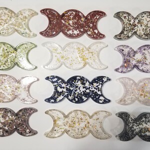 May include: Twelve triple moon resin trays with different glitter colors. The trays are clear and have a three-dimensional triple moon design. The glitter colors include gold, silver, green, purple, red, blue, and black.