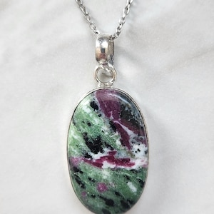 May include: An oval-shaped pendant necklace featuring a polished zoisite stone with prominent inclusions of deep magenta ruby and black hornblende. The stone is set in a simple silver-toned metal bezel and hangs from a matching silver-toned chain.
