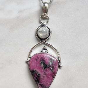 May include: A silver pendant necklace featuring a teardrop-shaped pink and black stone set in a polished silver frame. Above the main stone, a small round iridescent moonstone is set in a silver bezel, connected to a delicate silver chain.