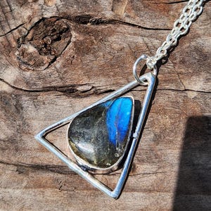 May include: A silver-toned necklace featuring a triangular metal frame pendant that holds a polished, irregular labradorite gemstone. The stone displays a dark grey base color with a prominent, vibrant blue flash across its surface. The pendant hangs from a matching silver-toned link chain.