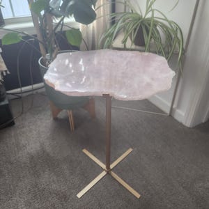 May include: A pink crystal table top with a gold metal base. The table top is shaped like a large, irregular oval. The base is a simple cross shape.
