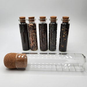 May include: Five small glass bottles with cork stoppers, each filled with different types of loose leaf tea. A sixth, empty bottle lies horizontally in front. The teas vary in color, from dark brown to light green.