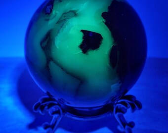 May include: A polished spherical stone featuring a vibrant, glowing green interior pattern with dark, opaque inclusions. The sphere is supported by a decorative, dark-colored metal stand with curved legs, illuminated under blue light.