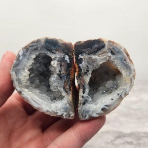 May include: Two halves of a geode, revealing a crystalline interior. The outer shell is a mix of brown, black, and white, while the inside sparkles with clear and gray crystals. The geode is held in a hand.