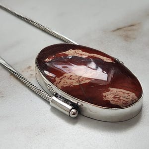 Peut inclure: Un collier en argent avec un pendentif ovale. Le pendentif pr&eacute;sente une pierre polie brun-rouge avec des motifs beiges et bruns. La pierre est sertie dans une lunette en argent et suspendue &agrave; une cha&icirc;ne en argent.