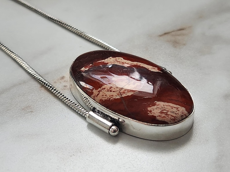 Peut inclure: Un collier en argent avec un pendentif ovale. Le pendentif pr&eacute;sente une pierre polie brun-rouge avec des motifs beiges et bruns. La pierre est sertie dans une lunette en argent et suspendue &agrave; une cha&icirc;ne en argent.