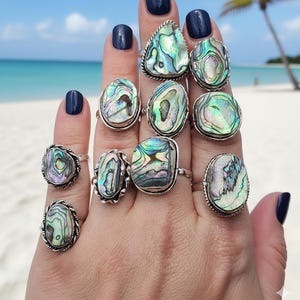 May include: A collection of silver rings with iridescent abalone shells in various shapes. The rings are displayed on a hand with dark blue nail polish, against a backdrop of a beach with blue water and a palm tree.