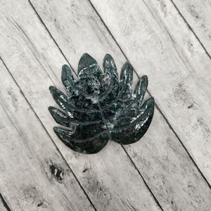 May include: A dark green leaf-shaped resin ornament with a glittery finish. The ornament is on a light wood background.