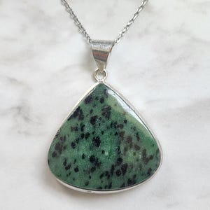 May include: A triangular pendant featuring a polished green stone with black speckled inclusions, set in a smooth silver-toned metal frame. The pendant hangs from a delicate silver-toned chain, showcasing a smooth, flat surface and a polished finish.