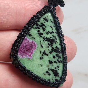 Peut inclure: Un pendentif triangulaire compos&eacute; d'une pierre polie verte avec des taches noires et une inclusion magenta distincte. La pierre est ench&acirc;ss&eacute;e dans un sertissage en cordon macram&eacute; noir avec une boucle nou&eacute;e au sommet pour le collier.