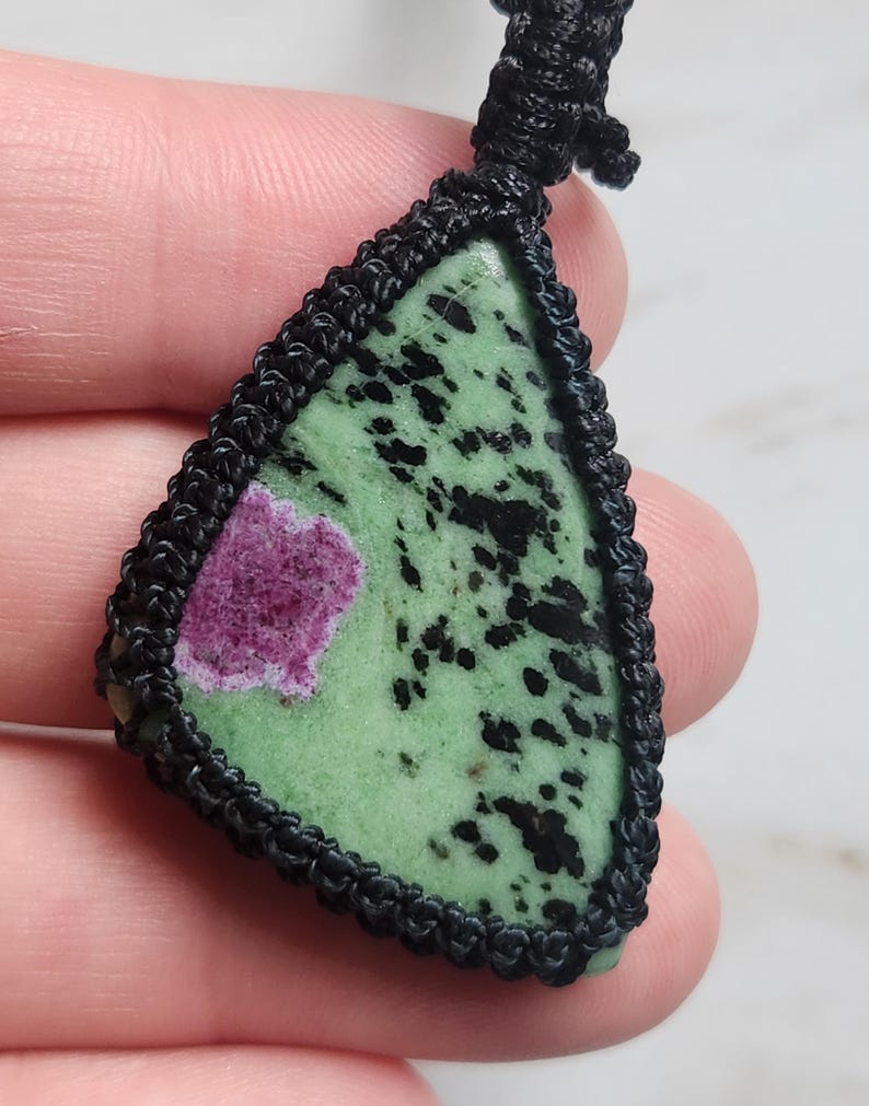 Peut inclure: Un pendentif triangulaire compos&eacute; d'une pierre polie verte avec des taches noires et une inclusion magenta distincte. La pierre est ench&acirc;ss&eacute;e dans un sertissage en cordon macram&eacute; noir avec une boucle nou&eacute;e au sommet pour le collier.