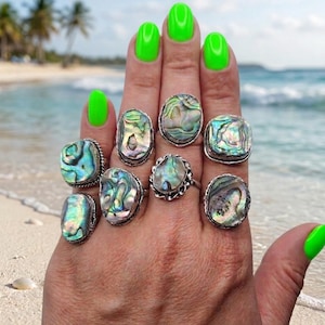 May include: A hand displays nine silver rings with iridescent abalone shells. The rings have various shapes, including oval and square, and are set on a beach with a turquoise ocean in the background. The nails are painted neon green.