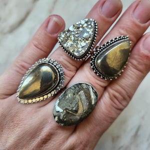 May include: Four rings featuring metallic pyrite stones set in silver-toned metal bands. Two rings have smooth, teardrop-shaped pyrite cabochons, one ring has a textured, crystalline pyrite surface, and one ring has a rough, faceted pyrite stone.