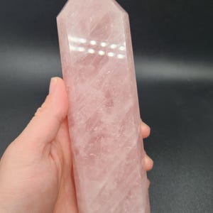May include: A polished rose quartz crystal obelisk. The pink crystal has a rectangular base and tapers to a point. The surface of the crystal has natural variations in color and texture. The obelisk is held in a hand against a dark background.