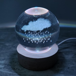 May include: A clear glass sphere with a cloud and rain design, illuminated by a soft light. The sphere sits on a wooden base, creating a calming, decorative piece. The design includes a rainbow effect.