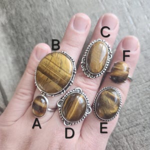 May include: Six silver rings with oval tiger's eye stones. The rings are labelled A through F.