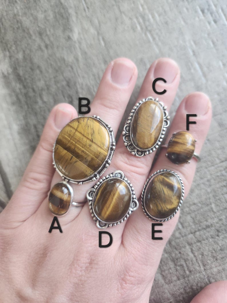 May include: Six silver rings with oval tiger's eye stones. The rings are labelled A through F.