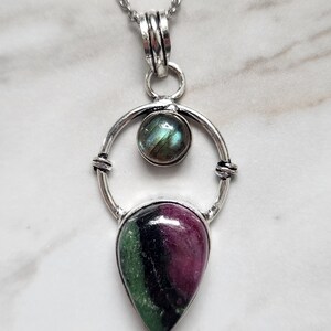 May include: A silver pendant necklace featuring a circular frame holding a small round iridescent labradorite stone at the top and a larger teardrop-shaped ruby zoisite stone with green, black, and magenta coloring at the bottom on a chain.