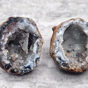 May include: Two halves of a geode, showcasing a hollow interior filled with sparkling crystals. The outer shell displays a mix of brown, black, and white bands. The geode halves are displayed on a light gray surface.