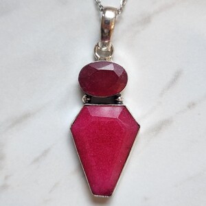May include: A silver pendant necklace featuring two faceted, deep pinkish-red stones set in metal. The top stone is oval-shaped, while the larger bottom stone is shield-shaped. The pendant hangs from a thin, silver-toned chain with a metallic finish.