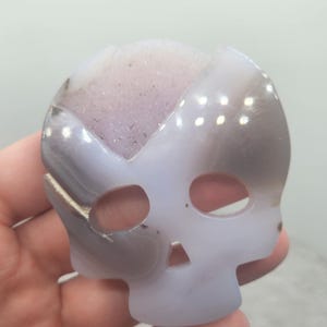 May include: A white agate skull with a purple and grey druzy interior. The skull is carved from a natural stone and has a smooth, polished finish.