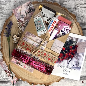 May include: A brown paper envelope decorated with pink and red sequins, burlap ribbon, and floral patterned paper. The envelope contains a card that says "Happy Valentine's Emma Love Tom".