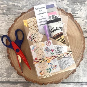 May include: A teacher appreciation gift set on a wooden slice. The set includes a small Galaxy instant chocolate, a purple pen, decorative lace, and cards with text that reads "Teachers are like bookmarks" and "You are the best teacher". Red and blue scissors are on the left.