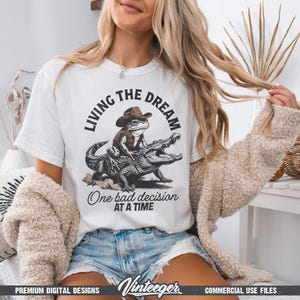 Puede incluir: Camiseta blanca con un dise&ntilde;o humor&iacute;stico de una rana con sombrero de vaquero montada en un caim&aacute;n. El texto dice "Living the Dream" arriba y "One bad decision at a time" abajo. Incluye el logotipo "Vinteeger".