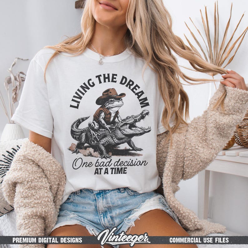 Puede incluir: Camiseta blanca con un dise&ntilde;o humor&iacute;stico de una rana con sombrero de vaquero montada en un caim&aacute;n. El texto dice "Living the Dream" arriba y "One bad decision at a time" abajo. Incluye el logotipo "Vinteeger".