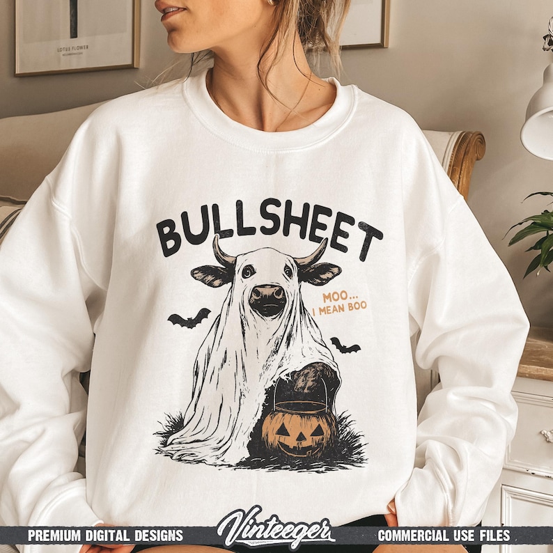 May include: White sweatshirt with a Halloween-themed graphic. The design features a cow dressed as a ghost, holding a jack-o'-lantern, with the text "BULLSHEET" above and "MOO... I MEAN BOO" below. Includes bat silhouettes.