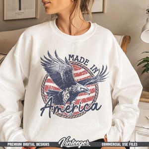 May include: White sweatshirt featuring a graphic of an eagle with outstretched wings, set against a red and white striped background. The text "MADE IN America" is displayed in a circular design. The sweatshirt has a crew neck.