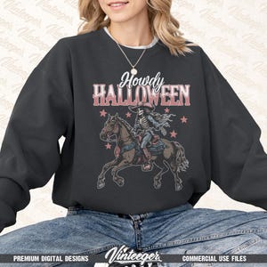 Op de afbeelding: Donkergrijze sweatshirt met de tekst "Howdy Halloween" boven een illustratie van een skelet te paard. Het ontwerp bevat roze sterren en een cowboyhoed. De sweatshirt is een premium digitaal ontwerp.