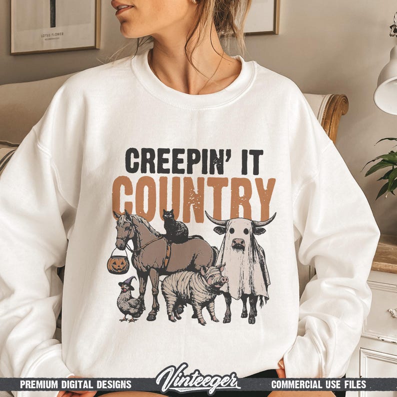 May include: White sweatshirt with the text "CREEPIN' IT COUNTRY" in black and brown. The graphic features a horse, cat, pig, chicken, and bull, all in Halloween costumes. The design has a vintage style.