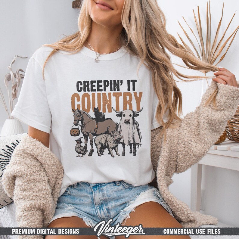 May include: White t-shirt with the text "CREEPIN' IT COUNTRY" in black and brown. The graphic features a horse, cow, and other animals in Halloween costumes. The shirt is a premium digital design.