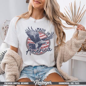May include: White t-shirt featuring a graphic of an eagle with the text "MADE IN America" in a circular design with red and white stripes. The shirt is a casual style.